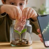 Kinderhände stecken einen 10-Euro-Schein in ein Glas mit Münzen und einem wachsenden Setzling, daneben zeigt ein Smartphone eine steigende Kurve als Symbol für die Frühstartrente.