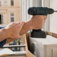 Hand setzt eine Bohrmaschine an die Außenwand eines Mietshauses, daneben ein neues Klima-Außengerät, eine zweite Hand stoppt den Einbau
