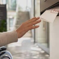 Frau im Rollstuhl greift nach einem Behördenbrief mit Fristvermerk in einem zu hoch angebrachten Briefkasten, im Hintergrund ein heller Behördenflur mit Treppe.