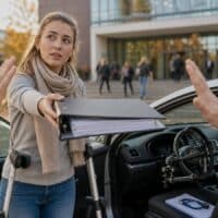 Studentin mit Gehstützen reicht neben einem umgebauten Auto einen Antragsordner, den zwei Hände hin- und herschieben, vor einem Hochschulgebäude