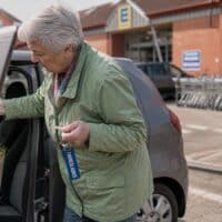 Ältere Frau mit Gehstock lädt auf einem Supermarktparkplatz Einkäufe in ihr Auto