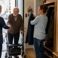 Pflegefachkraft wird im Hausflur von einer Frau und ihrem Vater mit Rollator begrüßt, an der Wand hängt ein Kalender mit dem eingekreisten Wort „TERMIN“