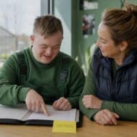 Junger Mann mit Trisomie 21 bespricht mit seiner Jobcoachin einen Arbeitsvertrag im Aufenthaltsraum eines Gartenbaubetriebs, auf der Mappe klebt ein Zettel mit dem Wort „VERTRAG“.