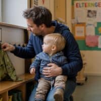 Vater hängt in einer Kita-Garderobe die Jacke seines etwa einjährigen Kindes an einen Haken, im Hintergrund eine Pinnwand mit dem Wort Elterngeld.
