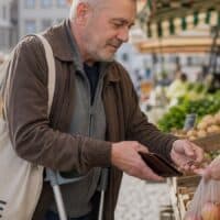 Mann Mitte fünfzig mit Unterarmgehstütze bezahlt an einem Gemüsestand auf einem Wochenmarkt mit Münzen, über seiner Schulter ein Stoffbeutel mit der Aufschrift „RENTE“.