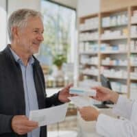 Älterer Mann mit Schwerbehinderung erhält in einer Apotheke Medikamente und eine Zuzahlungsübersicht für die Befreiung bei der Krankenkasse