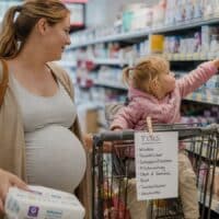 Schwangere Frau mit Kleinkind legt im Drogeriemarkt Windeln in den Einkaufswagen, am Griff hängt ein Einkaufszettel mit dem Wort PLUS.