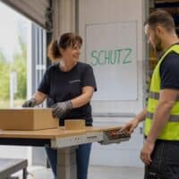 Lagermitarbeiterin probiert nach ihrer Rückkehr einen höhenverstellbaren Packtisch aus, während ein Kollege die Höhe anpasst; an der Schichttafel steht das Wort SCHUTZ.