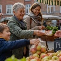Großmutter bezahlt mit ihrer erwachsenen Tochter und Enkelkind an einem Obststand auf dem Wochenmarkt, auf der Kreidetafel steht „PLUS“
