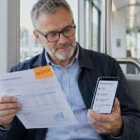 Mann um die 60 vergleicht in einer modernen Straßenbahn seine Rentenauskunft mit dem Smartphone, auf dem Dokument klebt ein oranger Zettel mit der Aufschrift „PRÜFEN“.
