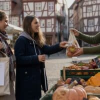 Junge Frau mit Blindenstock bezahlt an einem Marktstand, neben ihr ihre Assistentin mit einer Einkaufstasche.