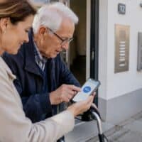 Tochter zeigt ihrem Vater mit Rollator vor dem Hauseingang auf dem Smartphone die neue Pflege-App, auf dem Display steht „NEU“.