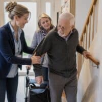 Pflegeberaterin zeigt einem älteren Mann bei einem Hausbesuch im Flur, wie er sich sicher am Treppenhandlauf hochzieht, die Tochter hält den Rollator.