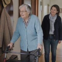 Ältere Frau geht mit ihrem Rollator durch den Flur ihrer Wohnung, während eine Gutachterin des Medizinischen Dienstes beim Hausbesuch aufmerksam zusieht