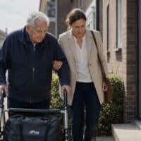 Berufstätige Tochter begleitet ihren Vater mit Rollator nach der Rückkehr aus dem Krankenhaus vom Auto zur Haustür
