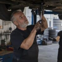 Kfz-Mechaniker Ende fünfzig arbeitet in einer Werkstatt unter einem angehobenen Auto, an der Wand eine Tafel mit der Aufschrift „Rente mit 65“.
