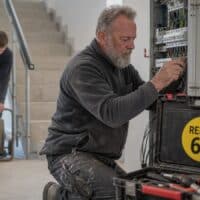 Elektriker Ende fünfzig montiert im Treppenhaus eines Neubaus einen Verteilerkasten, auf seinem Werkzeugkoffer klebt ein Aufkleber mit der Aufschrift „RENTE 65“