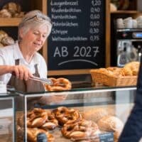 Rentnerin arbeitet im Minijob in einer Bäckerei und legt frische Brezeln in die Auslage, im Hintergrund eine Kreidetafel mit der Aufschrift AB 2027