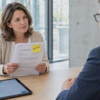 Hände halten in einer modernen Beratungsstelle einen ärztlichen Bericht mit gelber Haftnotiz „BEFUND“, daneben ein Tablet, der Hintergrund ist unscharf.