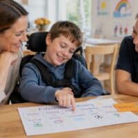 Mutter und Sohn im Rollstuhl sitzen mit einer Betreuerin am Esstisch und schauen auf einen Wochenplan mit Haftnotiz „BUDGET“