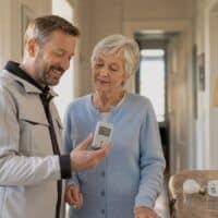 Energieberater zeigt einer Rentnerin im Wohnungsflur ein Strommessgerät, daneben LED-Lampe und Steckdosenleiste aus dem Stromspar-Check.