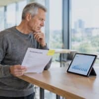 Älterer Mann in einem hellen Co-Working-Space hält einen Rentenbescheid mit gelber Haftnotiz und der Aufschrift FRIST, daneben ein Tablet mit Rentenübersicht.