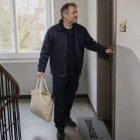 Mann mittleren Alters in Arbeitsjacke schließt im hellen Treppenhaus eines Mehrfamilienhauses seine Wohnungstür auf, auf der Fußmatte steht „Zuhause“.
