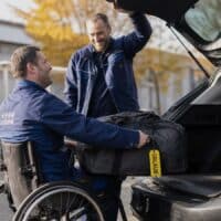 Mann im Rollstuhl lädt nach Feierabend eine Reisetasche in sein Auto, am Griff hängt ein Gepäckanhänger mit dem Wort Urlaub.